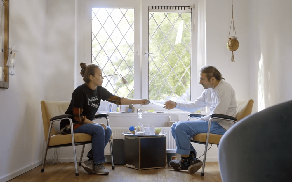 Zwei Menschen sitzen sich in einem hellen Raum auf zwei Stühlen vor einem Fenster gegenüber, eine Person reicht der anderen einen Flyer.